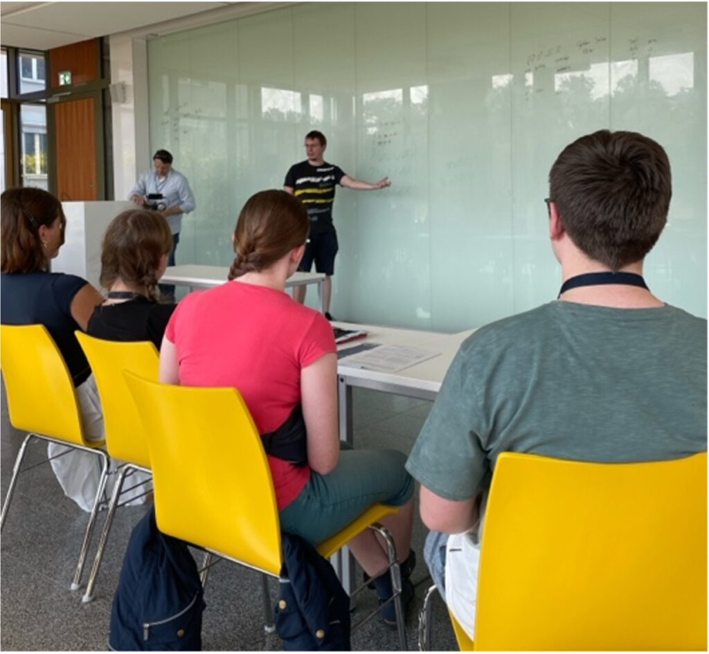 schüler sitzen in den MINT Sommerschulen mit dem rücken zur kamera und schauen auf eine tafel, an der ein lehrer mathematische formeln erklärt